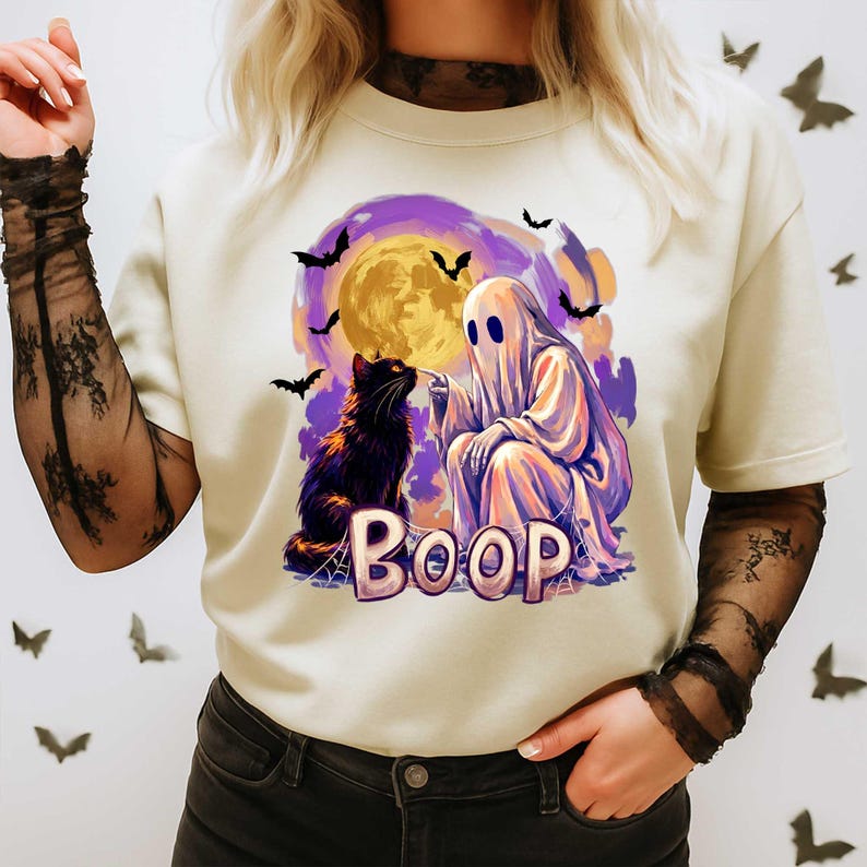 May include: A cream-colored t-shirt featuring a graphic of a white ghost touching the nose of a black cat. The design includes a large yellow moon, purple clouds, flying bats, and the word BOOP in large, light-colored, blocky letters.