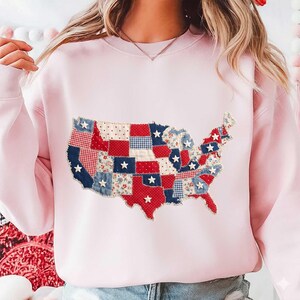May include: A light pink sweatshirt featuring a patchwork design of the United States map. The map is composed of red, white, and blue fabric squares with star accents, creating a patriotic theme.
