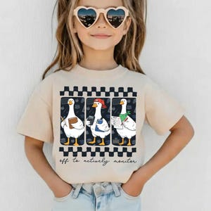 May include: Beige t-shirt with a graphic of three geese, each with school items. One goose holds a book, another a calculator, and the third a test paper. The shirt has a black and white checkered border and the text "off to actively monitor."