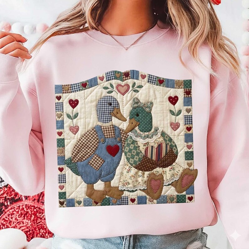May include: A light pink sweatshirt featuring a quilted design of two ducks. The ducks are adorned with heart-shaped accents and wear overalls and a dress. The design is framed with a patchwork border and heart motifs, creating a cozy, country aesthetic.
