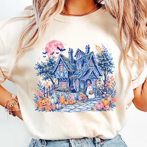 Pu&ograve; includere: Una t-shirt color crema con un'illustrazione grafica di una casa stregata blu di notte. Il design include una grande luna rosa, pipistrelli in volo, un gatto nero, un fantasma e zucche arancioni disposte attorno alla casa e un sentiero.