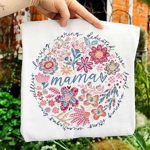 May include: White square pillow with a floral design. The design features a circular arrangement of flowers in pink, blue, and peach hues, surrounding the word "mama" in blue script. The words "loving, caring, dedicated, wise, loyal, inspiring, strong, brave, selfless" are written around the floral design.