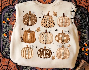 May include: A cream-colored crewneck sweatshirt featuring a graphic print of nine pumpkins arranged in a grid. Each pumpkin displays a unique pattern, including polka dots, leopard print, plaid, and floral designs, with football laces in the center.