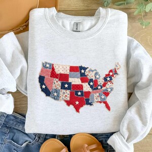 May include: A light gray sweatshirt featuring a patchwork design of the United States map. The map is composed of red, white, and blue fabric pieces with star accents. The sweatshirt is paired with blue jeans and sandals.