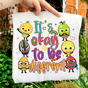 Op de afbeelding: Witte draagtas met de tekst "It's okay to be different" en kleurrijke cartoon fruit- en groentekarakters. Het ontwerp bevat een regenboog, sterren en harten.