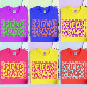 May include: Six colorful t-shirts with the text "FIELD DAY" in a retro font. The shirts are in various colors, including purple, blue, yellow, pink, and red. The phrase "LET THE GAMES BEGIN" is printed around the central text.
