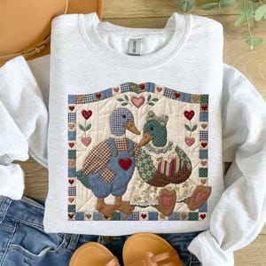 May include: A light gray sweatshirt featuring a quilted design of two ducks. The male duck wears blue overalls with a red heart, and the female duck wears a floral dress. The design includes hearts and floral accents.