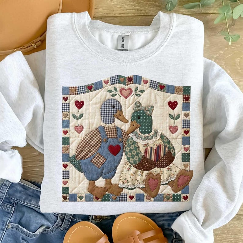 May include: A light gray sweatshirt featuring a quilted design of two ducks. The male duck wears blue overalls with a red heart, and the female duck wears a floral dress. The design includes hearts and floral accents.