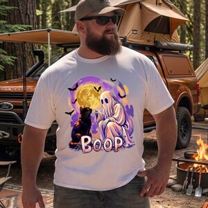 May include: A white short-sleeve t-shirt featuring a graphic print of a ghost touching the nose of a black cat. The design includes a yellow full moon, purple sky, flying bats, and the word BOOP in large, stylized lettering at the bottom.