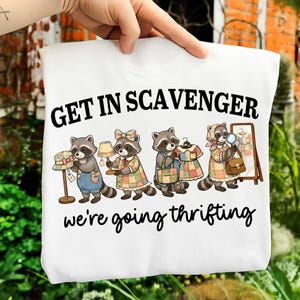 May include: White sweatshirt with a graphic of raccoons shopping. The text "GET IN SCAVENGER" is above the raccoons, and "we're going thrifting" is below. The raccoons are wearing outfits and holding items like a hat, lamp, and dresses.