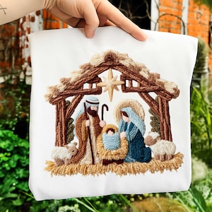 Könnte beinhalten: Weißer Stoff mit einer gestickten Krippenszene. Die Szene zeigt Maria, Josef, das Jesuskind und zwei Schafe unter einem rustikalen Holzbau mit einem Stern. Die Stickerei verwendet braune, blaue, beige und weiße Fäden.