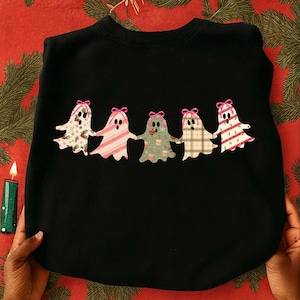 May include: A black crewneck sweatshirt featuring a graphic of five ghosts holding hands in a row. Each ghost has a pink bow on its head and a different holiday-themed pattern, including holly, candy cane stripes, ornaments, and plaid.