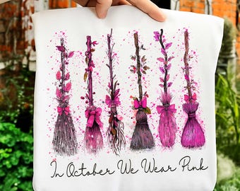 May include: White tote bag featuring watercolor illustrations of pink decorated witch brooms. The brooms are adorned with bows and foliage, set against a backdrop of pink paint splatters. The text "In October We Wear Pink" is written in a script font.