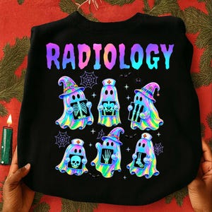 May include: A black crewneck sweatshirt featuring the word RADIOLOGY in a colorful gradient font. Below the text are six iridescent ghosts, some wearing witch hats or nurse caps, each holding a different medical X-ray image of human bones.