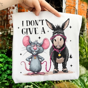 Puede incluir: Bolso tote blanco con una ilustraci&oacute;n de dibujos animados de un rat&oacute;n gris y un burro negro. El rat&oacute;n tiene orejas rosas y una cola rosa. El burro lleva un cabestro rosa. El texto "I DON'T GIVE A" est&aacute; impreso en la parte superior.