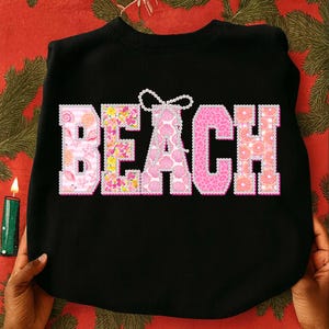 Puede incluir: Sudadera negra con la palabra "BEACH" en letras rosas. Las letras están decoradas con patrones de conchas marinas, flores y frutas. La sudadera es sostenida por dos manos.