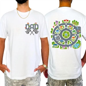 Könnte beinhalten: Weißes T-Shirt mit farbenfroher Grafik. Auf der Vorderseite steht das Wort "DAD" in einem Straßen-Design mit karierten Flaggen. Auf der Rückseite ist eine detaillierte Illustration einer Stadt mit Straßen, Häusern, Bäumen und einem Teich zu sehen.