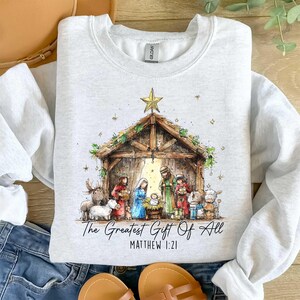 May include: Light gray sweatshirt featuring a Nativity scene with the text "The Greatest Gift Of All" and "Matthew 1:21". The graphic depicts the Holy Family, animals, and a star. The sweatshirt is a Gildan brand.