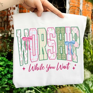 May include: White sweatshirt with the word "WORSHIP" in a patchwork design with pink and blue bows. The phrase "While You Wait" is written below in pink script, with pink star accents.