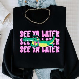 May include: Black sweatshirt with the phrase "SEE YA LATER" in pink, repeated three times. A cartoon alligator wearing sunglasses and a pink bow tie is in the center. A gold chain necklace is draped on top.