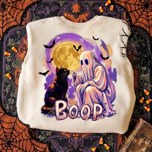 May include: A cream-colored crewneck sweatshirt featuring a graphic print of a white ghost booping the nose of a black cat. The background shows a large yellow moon, purple clouds, and black bats. The word BOOP is printed in large letters.