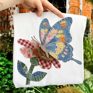 Op de afbeelding: Wit vierkant kussen met een kleurrijke vlinder- en bloemenapplicatie. De vlinder heeft blauwe spijkerbroekvleugels met roze bloemen- en gele ruitjesstofvlekken. De bloem heeft rood-witte ruitjesblaadjes en een bruin midden.