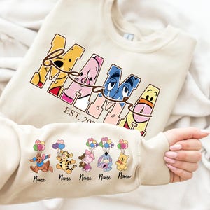 Könnte beinhalten: Cremefarbenes Sweatshirt mit dem Wort "MAMA" in einer farbenfrohen Cartoon-Schriftart, wobei die "A"s als Winnie-the-Pooh-Charaktere gestaltet sind. Der Ärmel zeigt Zeichentrickfiguren mit Luftballons. Der Text "EST. 20--" ist ebenfalls sichtbar.