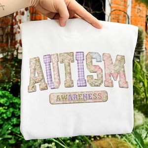以下が含まれることがあります： 花柄とギンガムチェックのパッチワークデザインの「AUTISM」という文字が入った白いスウェットシャツ。「AUTISM」の下には、花柄のデザインで「AWARENESS」という文字が入っています。スウェットシャツは手で持たれています。