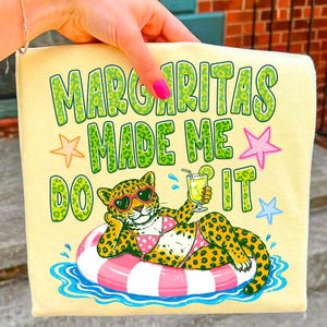 May include: A light yellow beach towel with the text "Margaritas Made Me Do It" in green. The towel features a cartoon leopard wearing sunglasses and a bikini, floating on a pink and white inner tube, holding a cocktail.