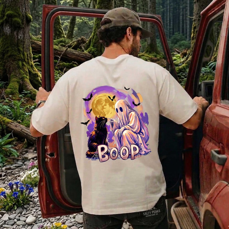 May include: A cream-colored t-shirt featuring a graphic on the back of a white ghost booping the nose of a black cat. The design includes a large yellow full moon, purple clouds, and black bats. The word BOOP is printed in large letters below.