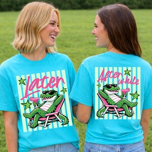 May include: Two turquoise t-shirts with a cartoon alligator wearing sunglasses and holding a pink cocktail. The front of the shirt says "Later" and the back says "After a while". The alligator is lounging in a pink and white striped chair.
