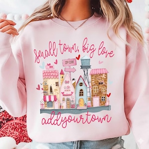 May include: Pale pink sweatshirt featuring a small town graphic. The design includes colourful buildings, a water tower, and the phrases "small town, big love" and "add your town" in cursive. Hearts and a zip code sign are also included.
