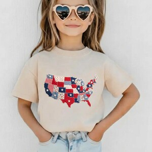 May include: A beige t-shirt featuring a patchwork design of the United States map in red, white, and blue. The map includes stars and various patterns. The shirt is short-sleeved and worn by a person.