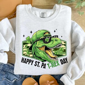 May include: A light grey sweatshirt with a green T-Rex wearing sunglasses and a hat, with the text "HAPPY ST. PA T-REX DAY". The design includes shamrocks and a camouflage background.