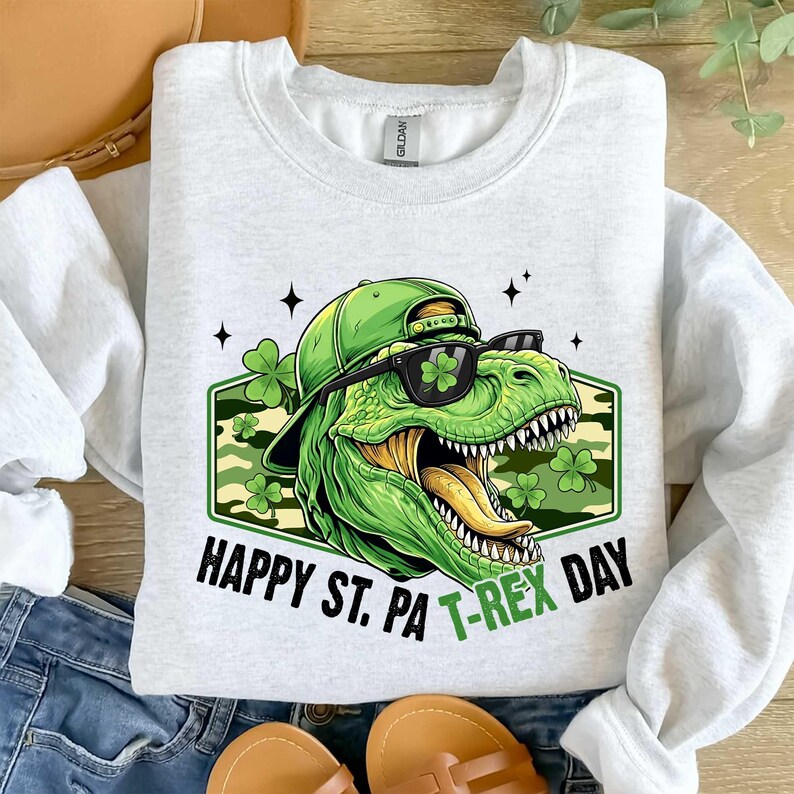 May include: A light grey sweatshirt with a green T-Rex wearing sunglasses and a hat, with the text "HAPPY ST. PA T-REX DAY". The design includes shamrocks and a camouflage background.