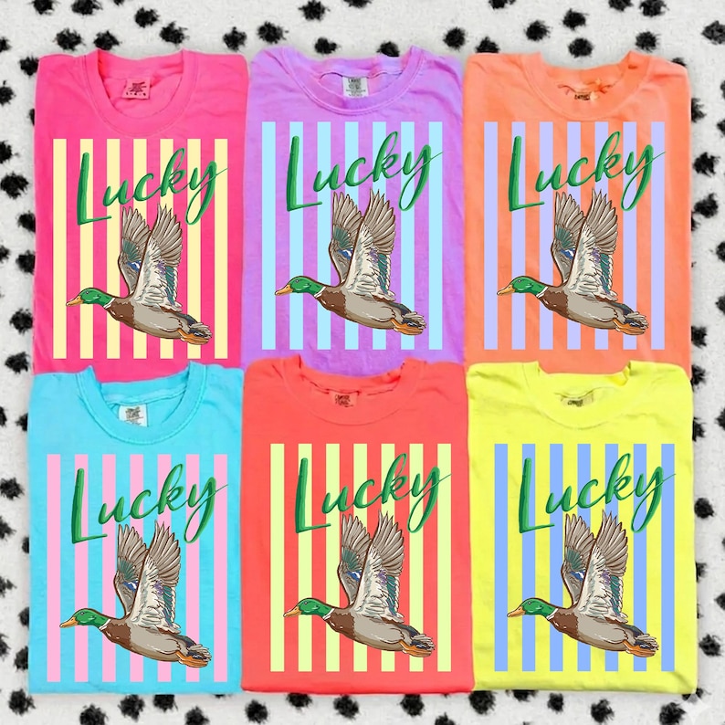 May include: Six colorful t-shirts in pink, purple, orange, blue, red, and yellow. Each shirt features a vertical striped design with the word "Lucky" in green script and a graphic of a flying duck.