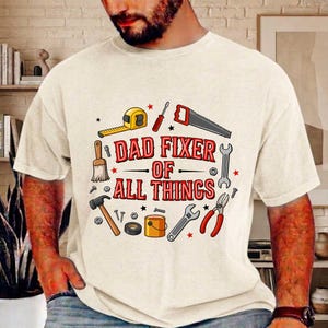 May include: Cream-colored t-shirt with a graphic of tools and the text "DAD FIXER OF ALL THINGS" in red and black. The tools include a tape measure, saw, wrench, and hammer.