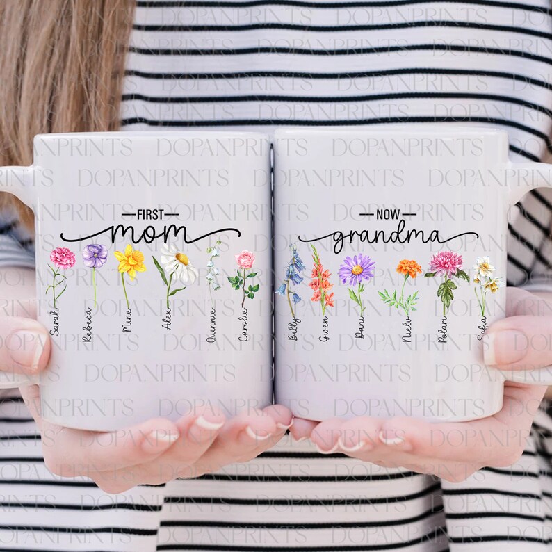 Puede incluir: Dos tazas de cer&aacute;mica blanca con asas. La taza de la izquierda tiene el texto "First Mom" y la taza de la derecha tiene el texto "Now Grandma". Ambas tazas tienen una l&iacute;nea de flores con nombres escritos debajo de cada flor.