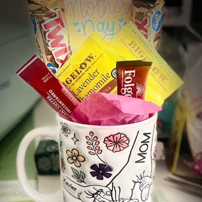 Op de afbeelding: Een witte keramische mok met een bloemenpatroon en het woord "MOM" erop gedrukt. De mok is gevuld met roze tissuepapier en verschillende items, waaronder een doos instantkoffie, een doos thee, een doos warme chocolademelk en een chocolade reep.