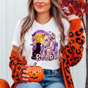 May include: A white short-sleeve t-shirt featuring a graphic print of a ghost and a black cat sitting under a yellow moon with purple clouds and bats. The text BOOP is printed in large letters below the ghost and cat. The shirt is worn with an orange leopard print cardigan.