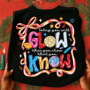May include: Black sweatshirt with the text "Today you will GLOW when you show what you KNOW" in colorful, playful lettering. The design includes a globe, stars, and school-themed illustrations.