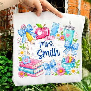 May include: A white t-shirt featuring a colorful graphic of school supplies including a notebook page with the text Mrs. Smith, a red apple, a yellow pencil, a blue ruler, stacked books, a teal tumbler, and blue gingham bows with flowers.