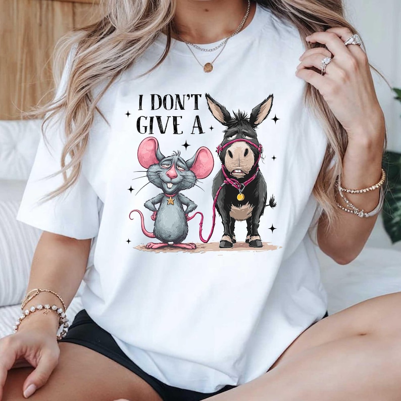 Puede incluir: Camiseta blanca con el texto "I DON'T GIVE A." y una ilustraci&oacute;n de dibujos animados de un rat&oacute;n gris y un burro negro. El rat&oacute;n tiene orejas rosas y el burro lleva un cabestro rosa.