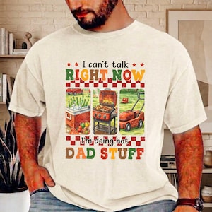 Puede incluir: Camiseta color crema con el texto "I can't talk RIGHT NOW I'm doing hot DAD STUFF." El gráfico incluye ilustraciones de una nevera portátil, una parrilla y un cortacésped. La camiseta es de diseño casual y de manga corta.