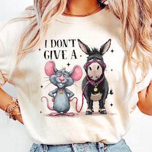 Puede incluir: Camiseta color crema con un rat&oacute;n y un burro de dibujos animados con el texto "I DON'T GIVE A." El rat&oacute;n es gris con orejas rosas y una estrella dorada. El burro es negro con un cabestro y una correa rosas.