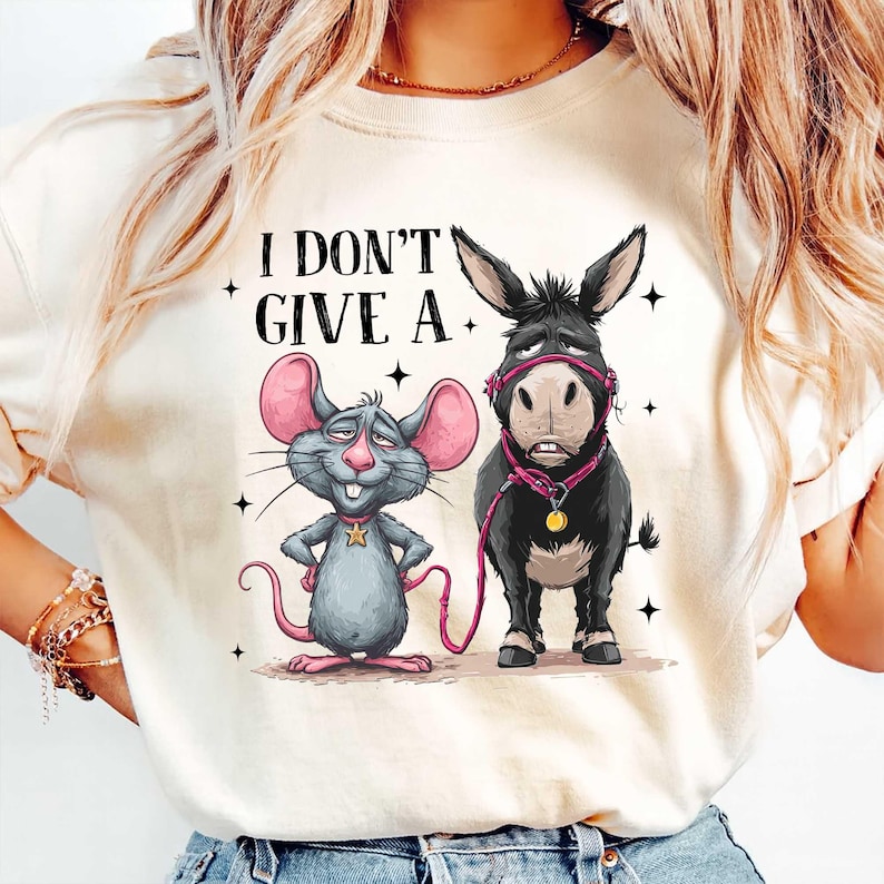 Puede incluir: Camiseta color crema con un rat&oacute;n y un burro de dibujos animados con el texto "I DON'T GIVE A." El rat&oacute;n es gris con orejas rosas y una estrella dorada. El burro es negro con un cabestro y una correa rosas.