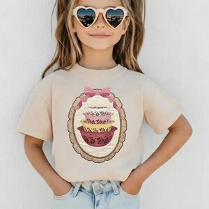 May include: A beige t-shirt featuring a graphic of stacked, vintage-style mixing bowls in pink, yellow, and white, framed by a pink bow and an oval border. The shirt is worn by a person wearing heart-shaped sunglasses.