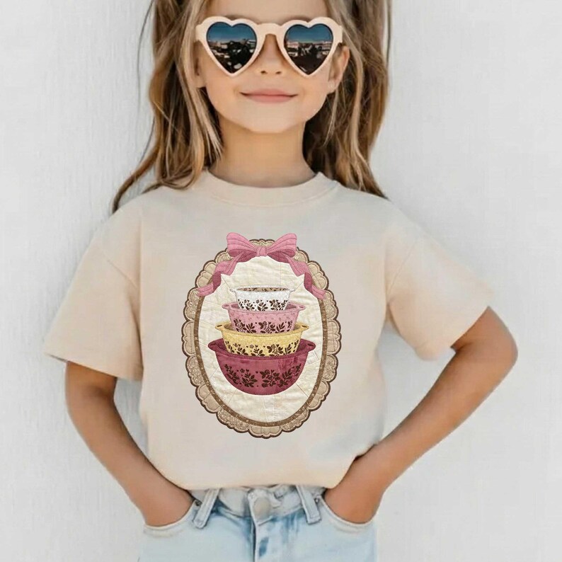 May include: A beige t-shirt featuring a graphic of stacked, vintage-style mixing bowls in pink, yellow, and white, framed by a pink bow and an oval border. The shirt is worn by a person wearing heart-shaped sunglasses.