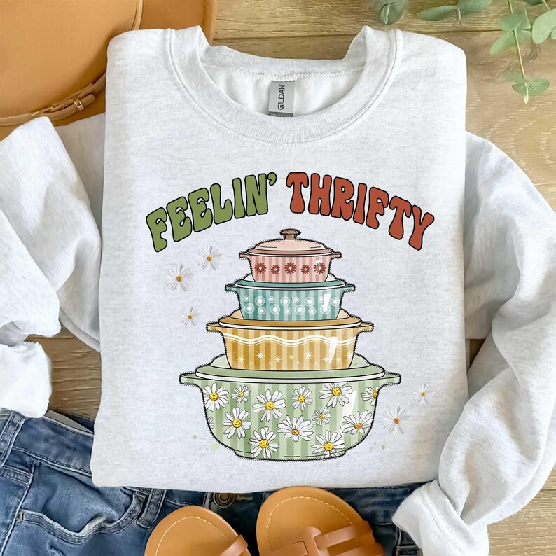 May include: A light gray sweatshirt with the text "FEELIN' THRIFTY" in green and orange. The graphic features a stack of colorful vintage casserole dishes with floral and striped patterns. Daisies are scattered around the design.