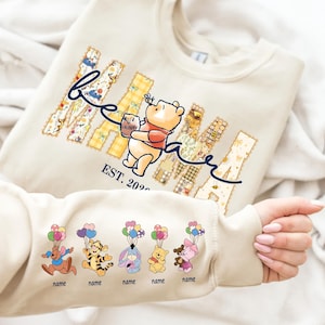 May include: Cream-colored sweatshirt featuring a Winnie the Pooh design. The word "Mama" is displayed in patchwork letters, with "be" in cursive. Winnie the Pooh is holding a honey pot. The sleeve displays cartoon characters with balloons. "EST. 2020" is printed below.